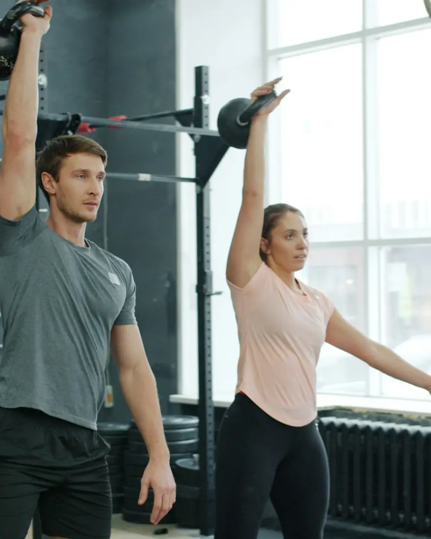 A coach and a member swing kettlebells overhead in a bright gym.