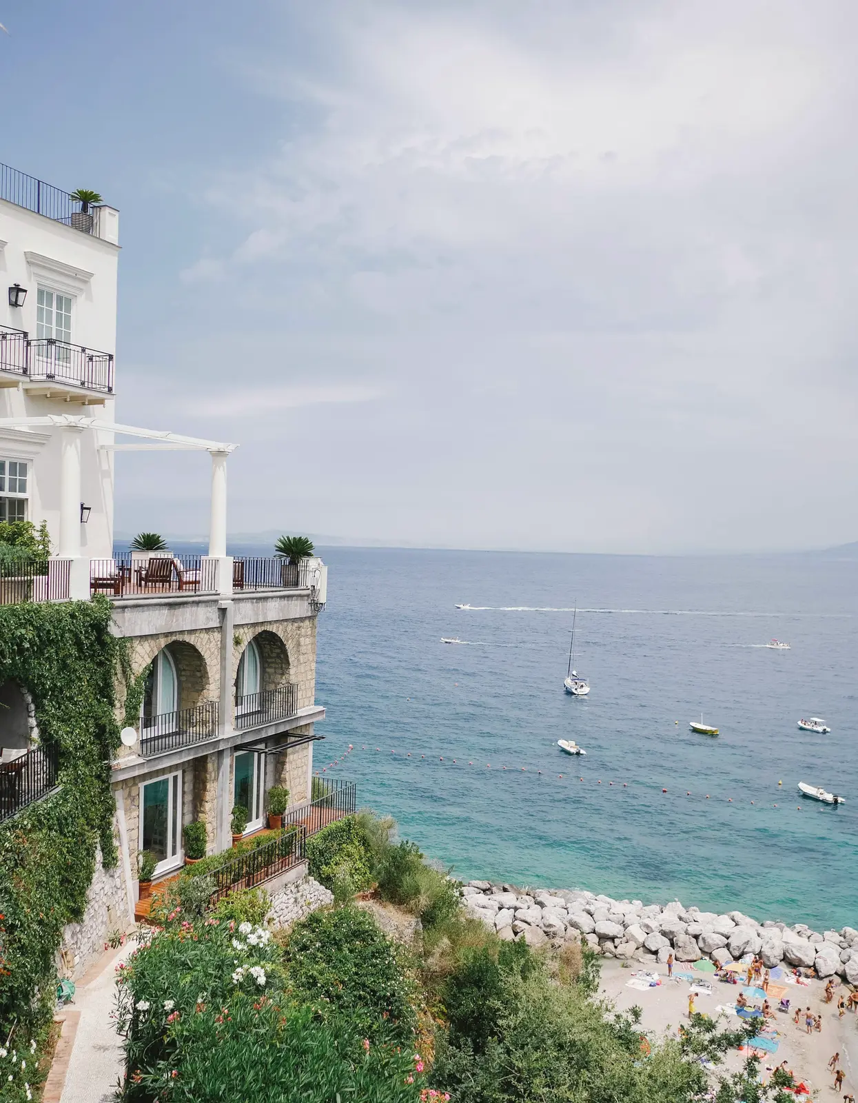 Villas and gardens stepping down a green cliff to a pebble beach and boats on a blue sea