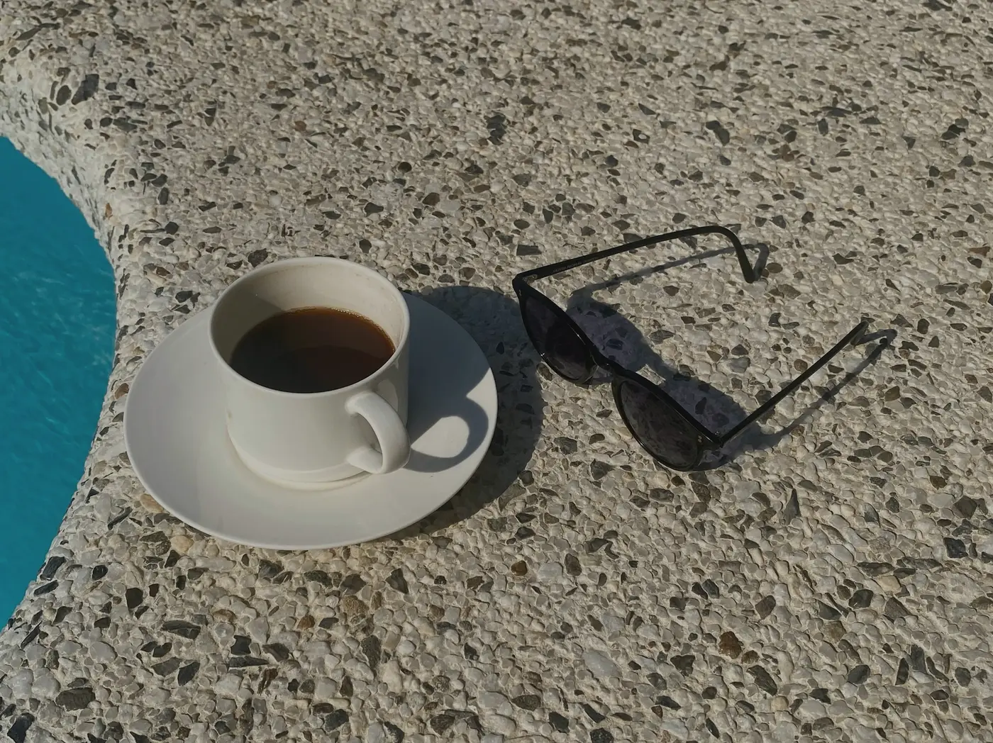A cup of black coffee and sunglasses on the stone edge of a pool