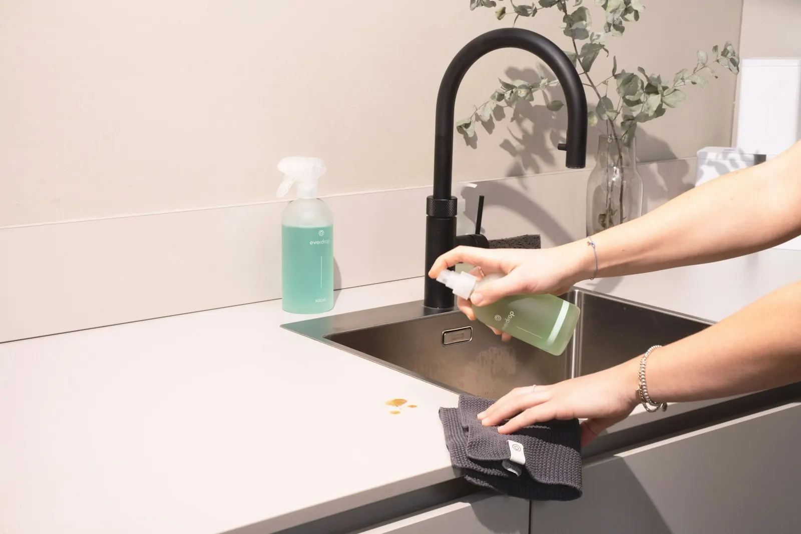 A cleaner's hands wiping down a dark kitchen sink with a cloth and a glass spray bottle