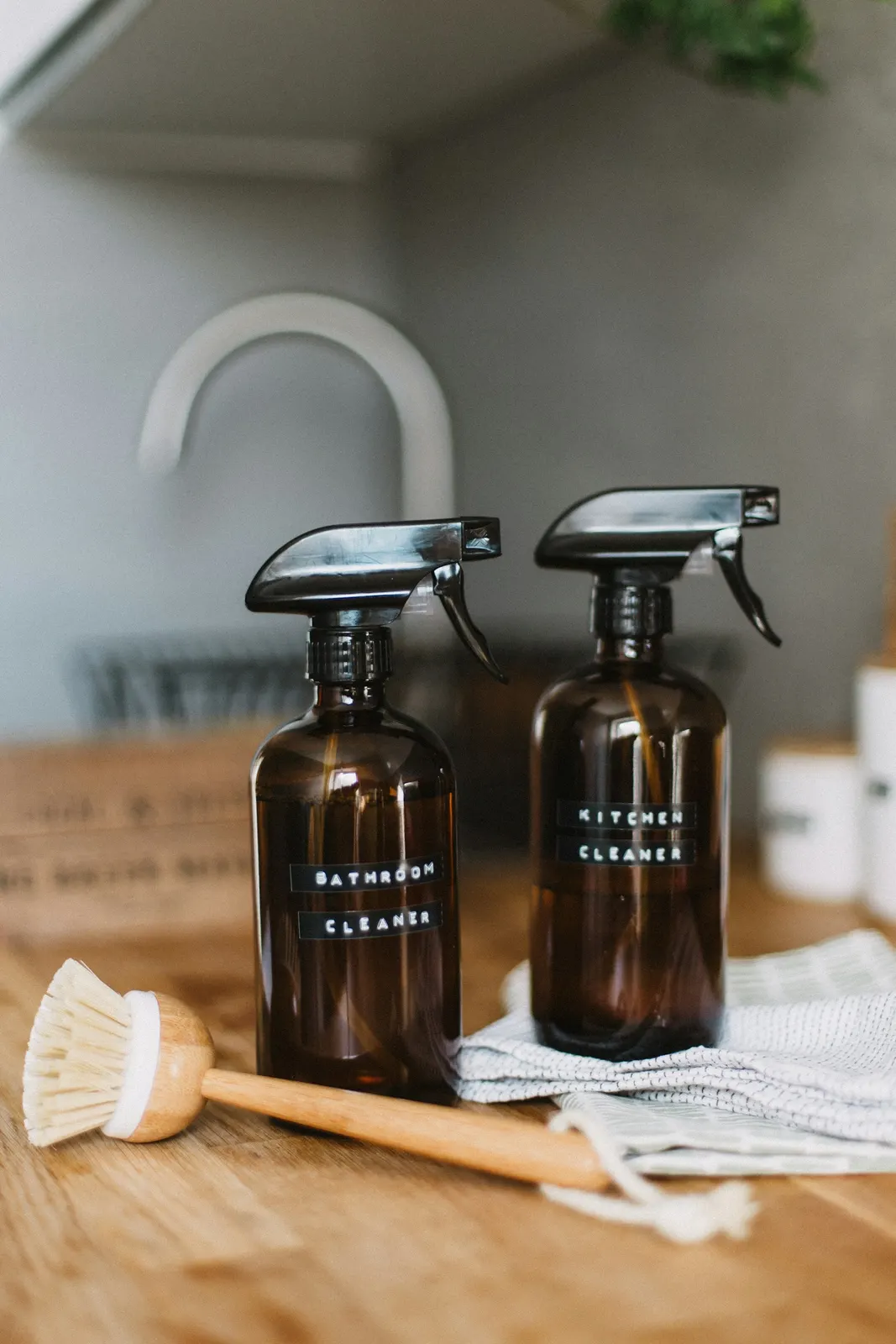 Two amber glass spray bottles of plant-based cleaner and a wooden scrub brush on a counter