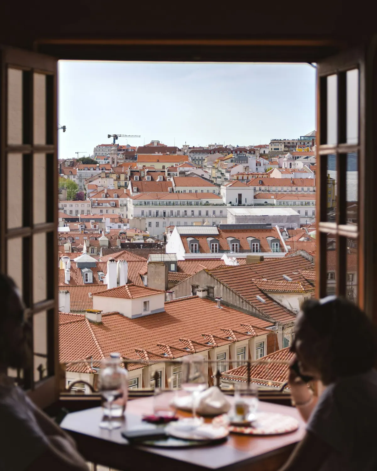 Two people at a dining table beside an open window overlooking the terracotta rooftops of Lisbon