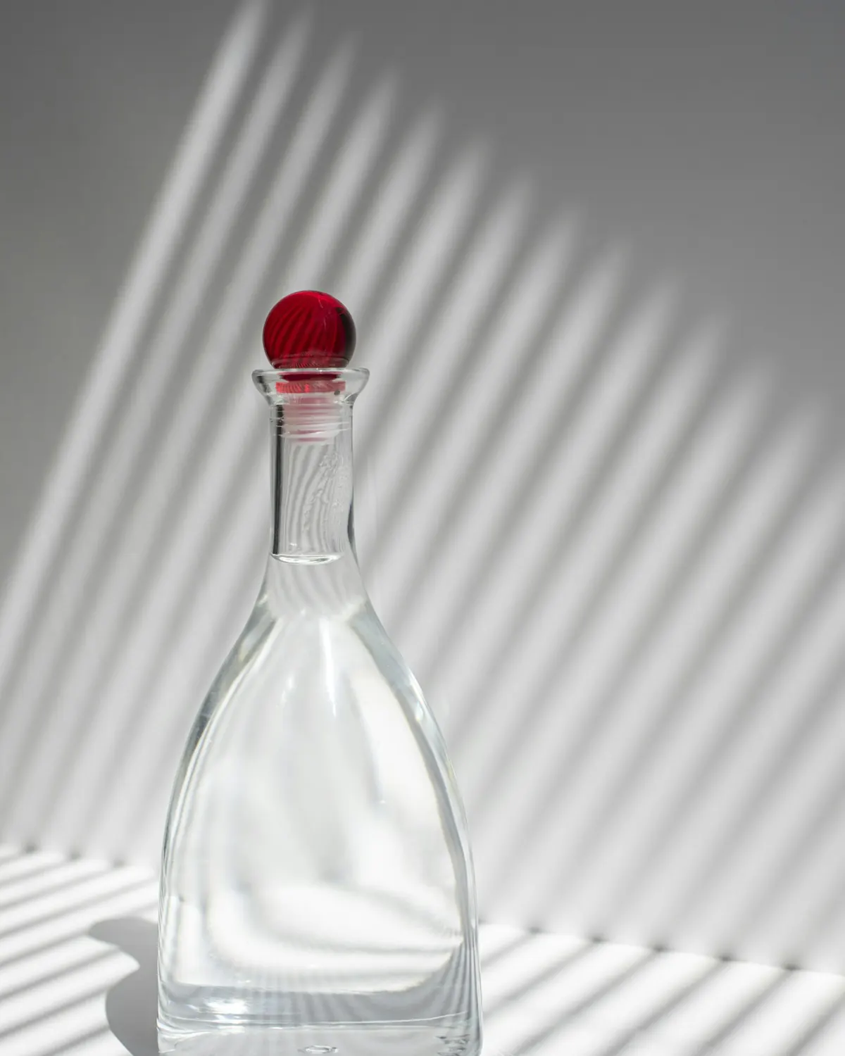 A clear glass bottle with a red stopper on a white surface, striped with window shadows