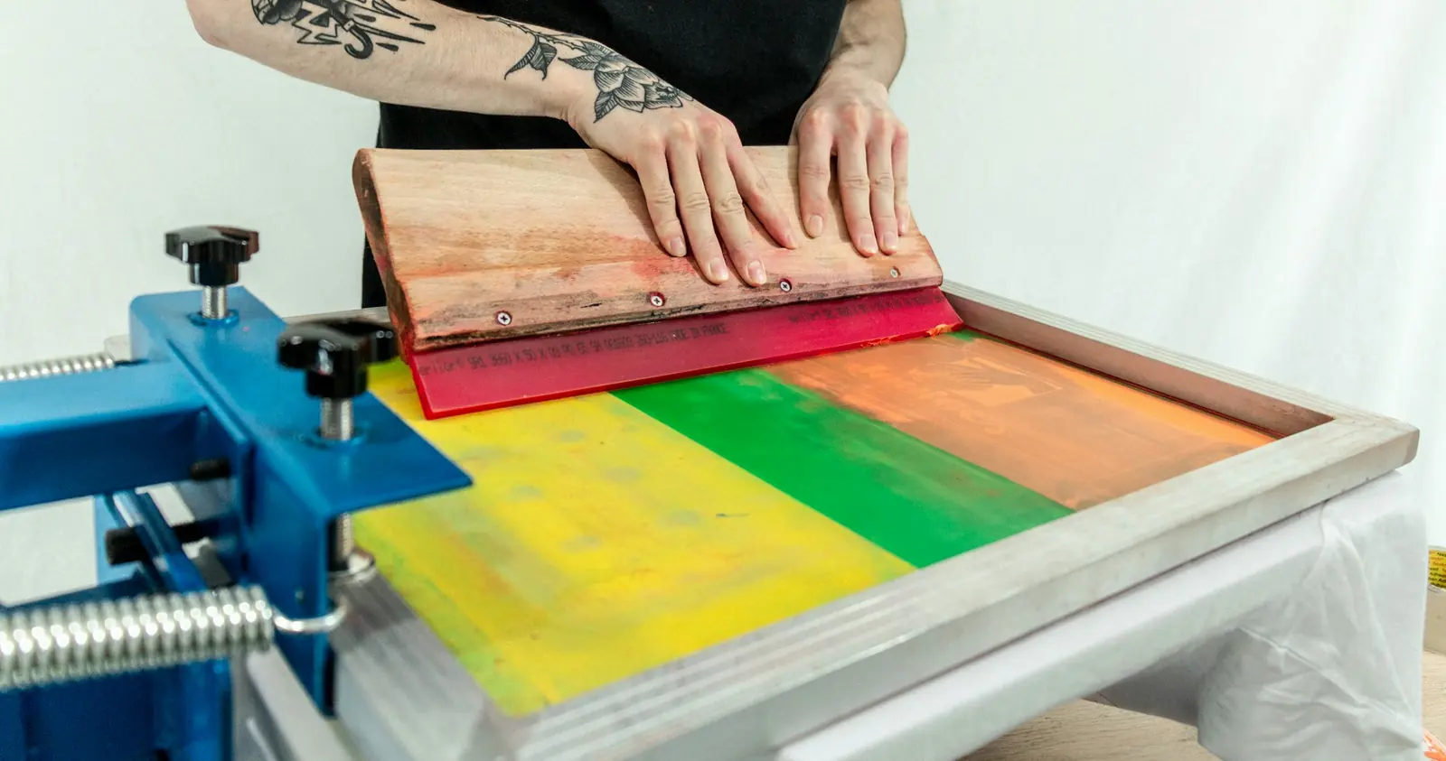 A printer pulls a squeegee across a screen, laying down a yellow, green and orange split-fountain print