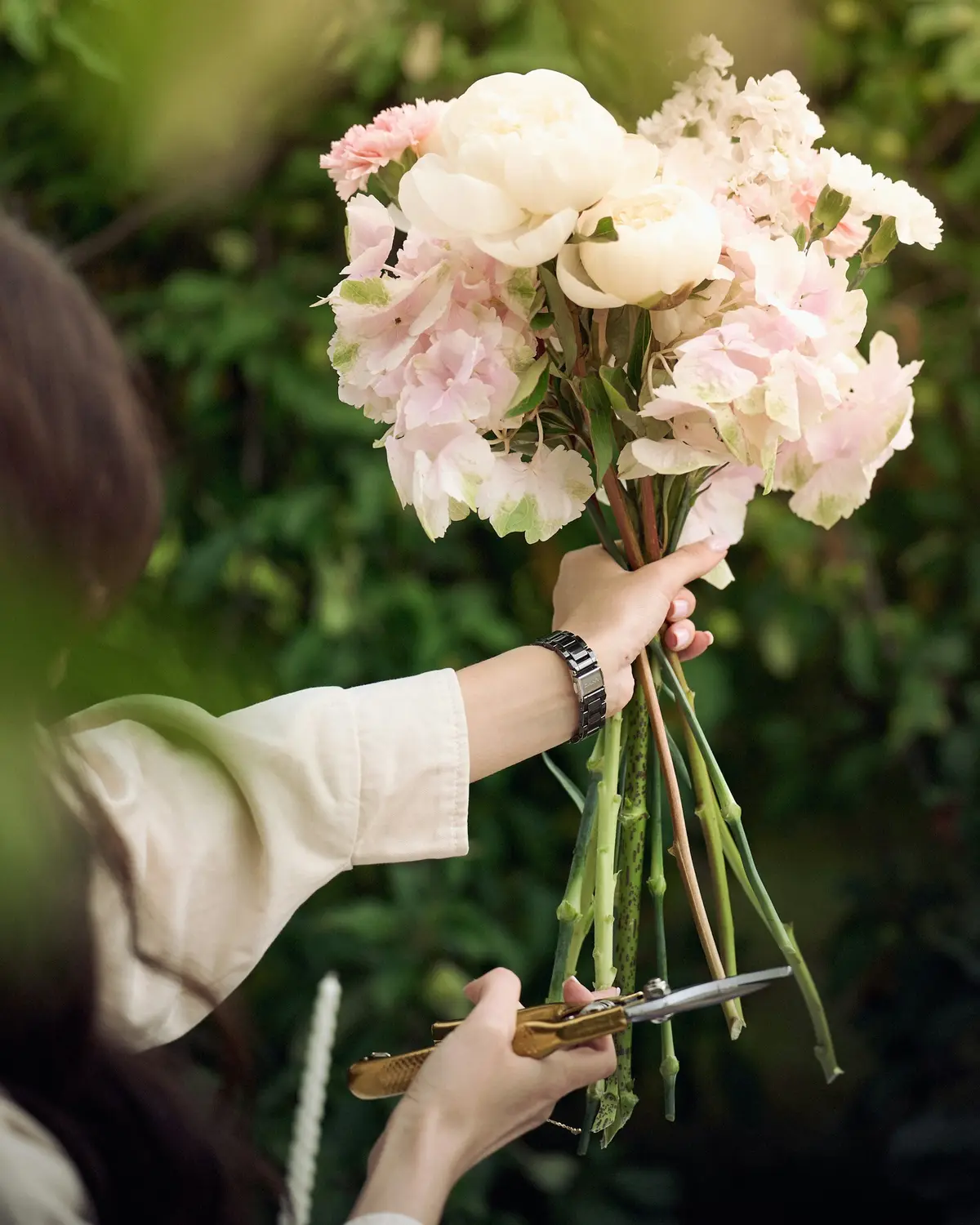 A hand holding up a loose bouquet of white garden roses, peonies and blush hydrangea while the stems are trimmed
