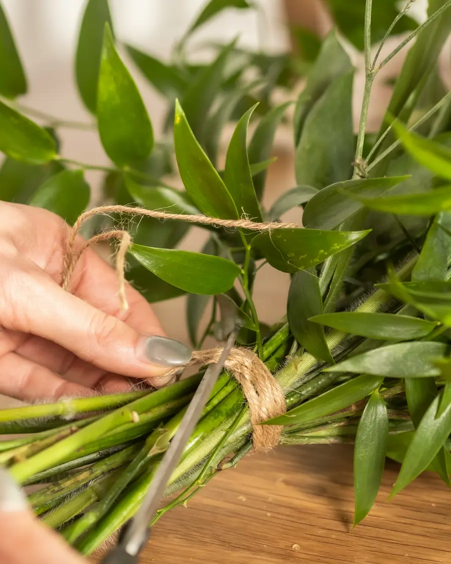 Hands tying a bundle of green stems with twine on a wooden bench