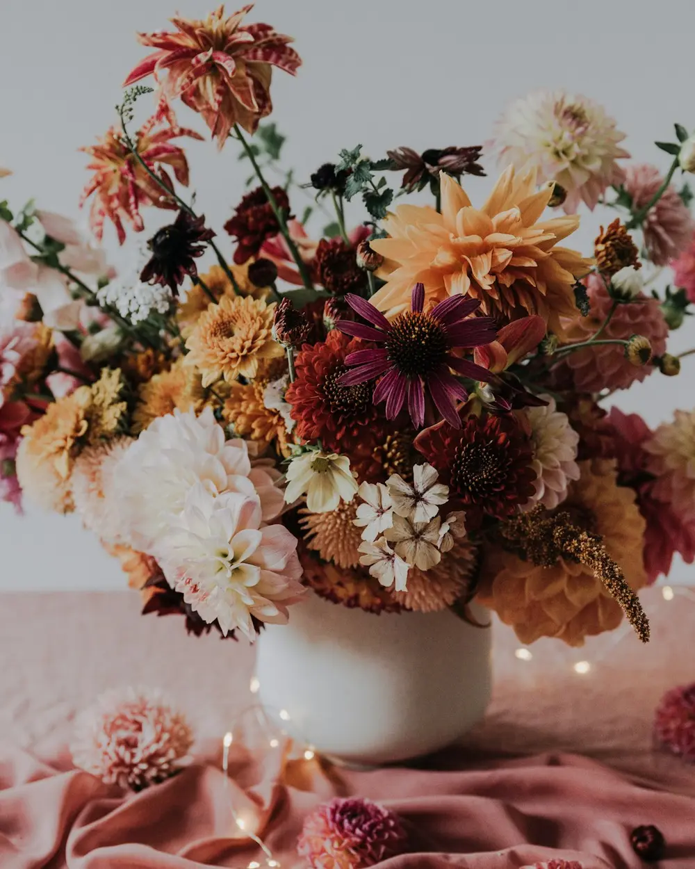 A loose low arrangement of rust and blush dahlias in a white footed bowl on a pink cloth