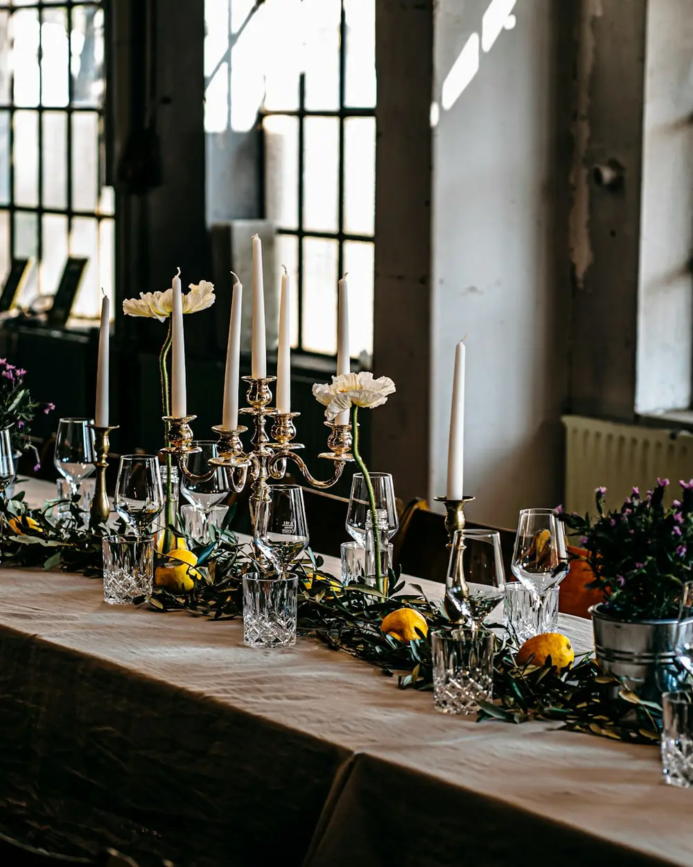A long candlelit table in an industrial loft with tall windows and low flowers along its length