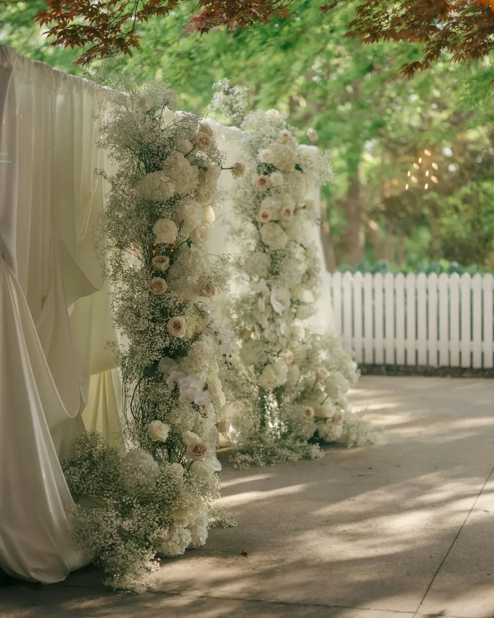Two tall pillars of white roses and baby's breath under trees at a garden ceremony
