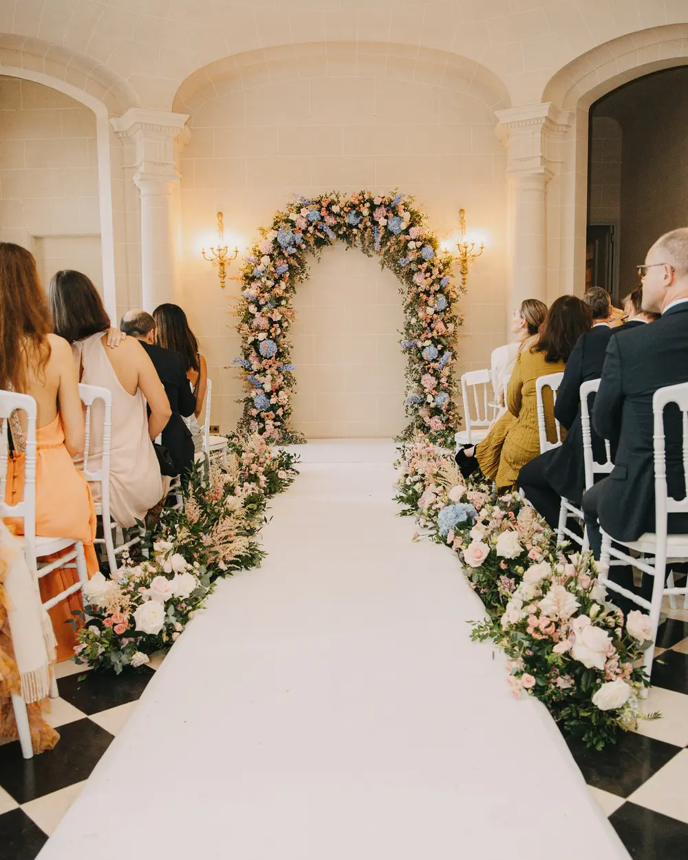 A white aisle through a chandeliered hall to an arch of blush and ivory flowers, guests seated either side