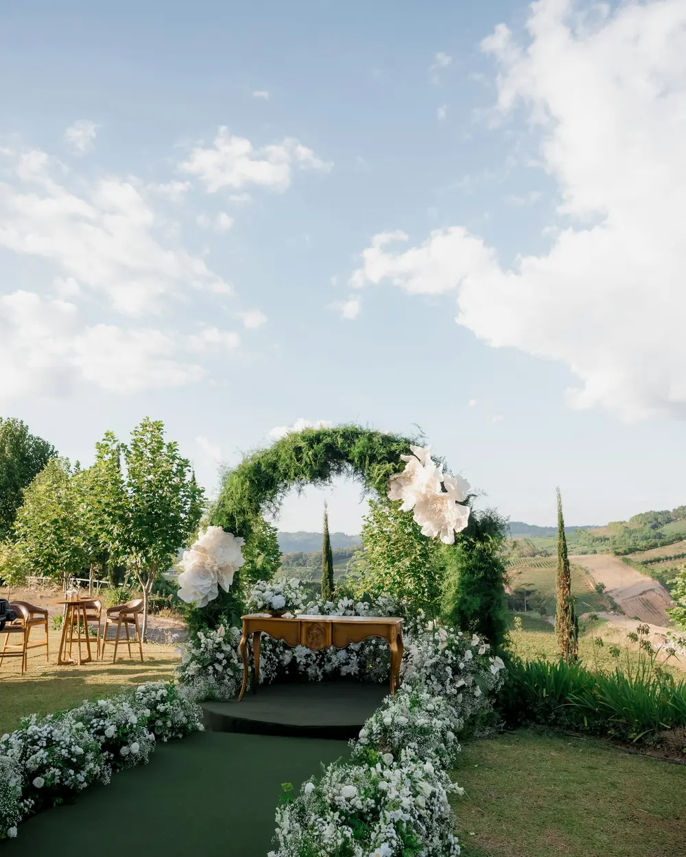 A green hoop arch and white flower-lined aisle on a lawn under a wide summer sky
