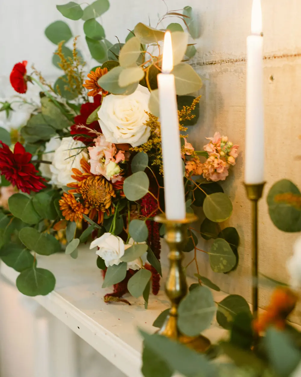 Red, orange and white flowers with eucalyptus around lit taper candles on a mantel