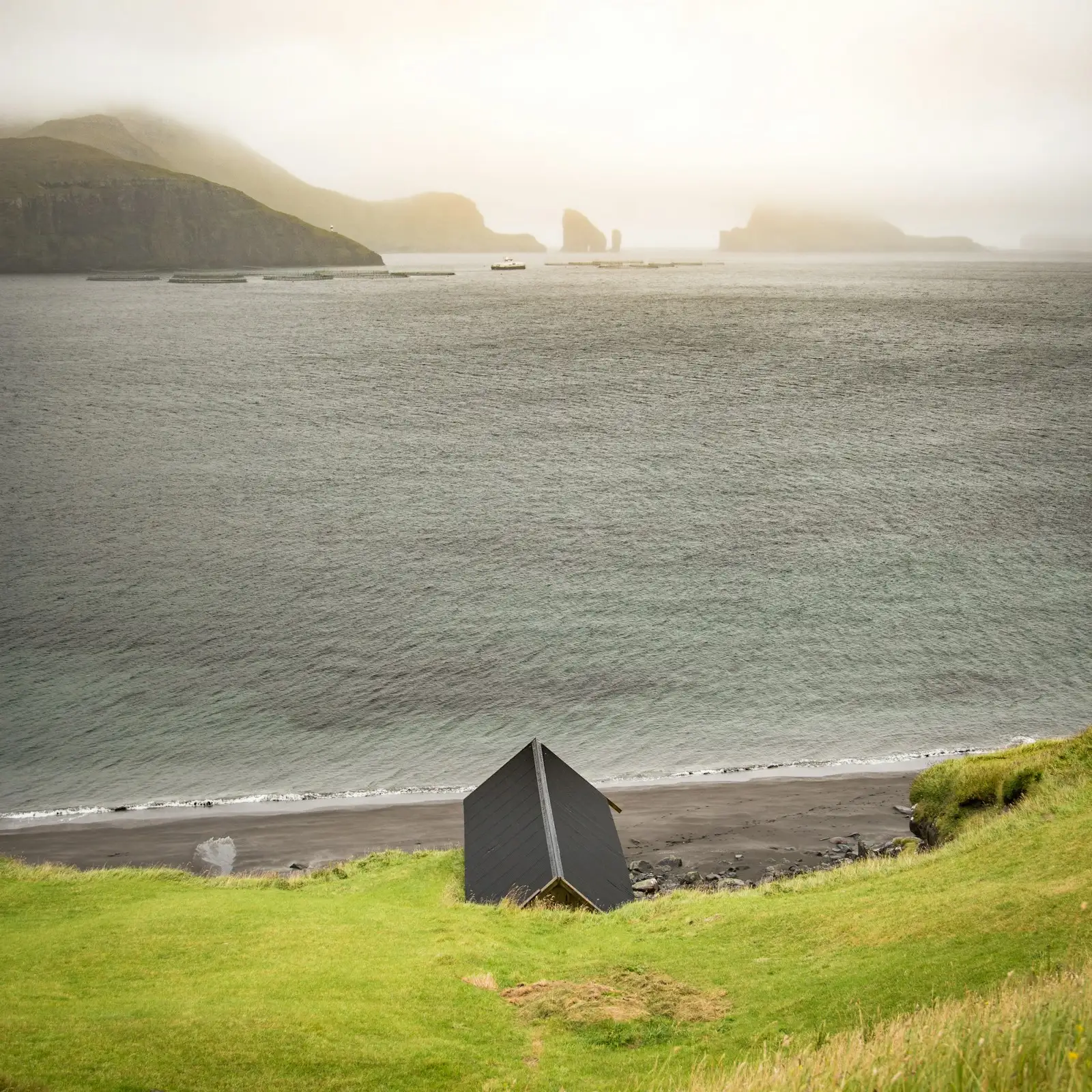 A small black-clad shelter on a green headland above a grey sea loch, hills beyond.