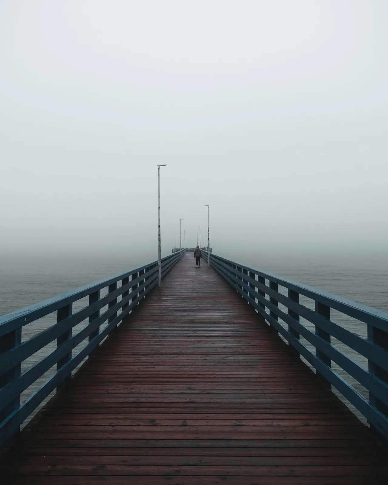 A wooden jetty with dark railings running out into a blank grey sea, two small figures at its far end.