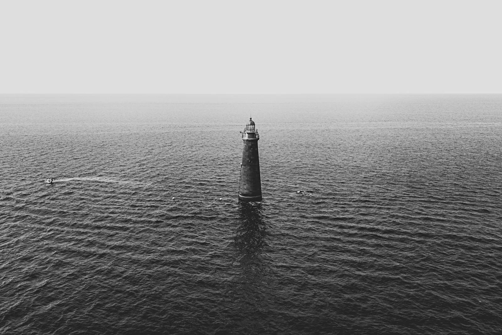 A slim lighthouse tower standing alone in rippled grey water under a white sky.