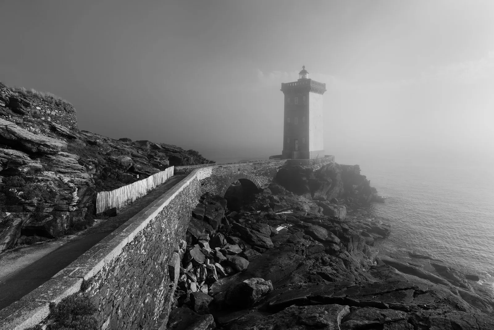 Black-and-white view of a lighthouse on a rocky headland, a stone wall curving towards it through fog.
