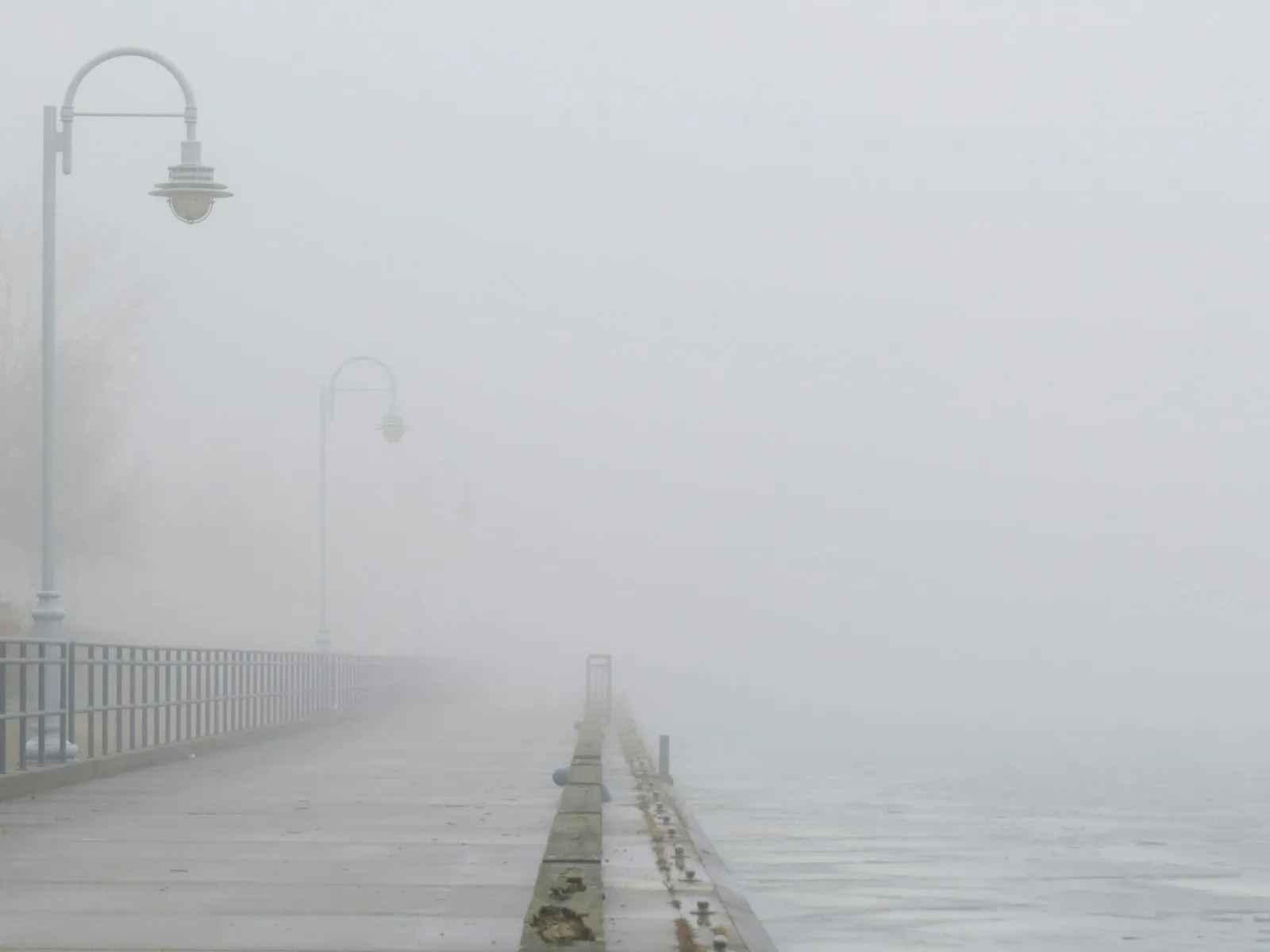 A curved lamp post and pier railing fading into white fog over flat water.