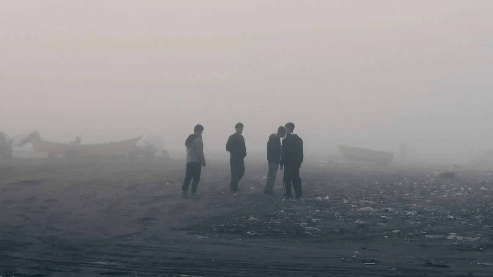 Four men standing and talking on a grey beach, half erased by fog.
