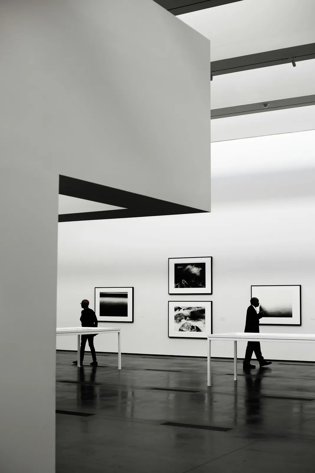 Two visitors walking past framed black-and-white photographs in a white, top-lit gallery with a polished dark floor.