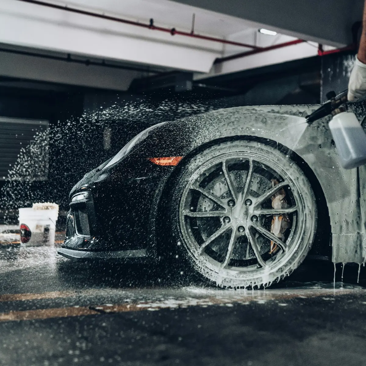 A dark sports car covered in wash foam, a detailer's sprayer beside the front wheel