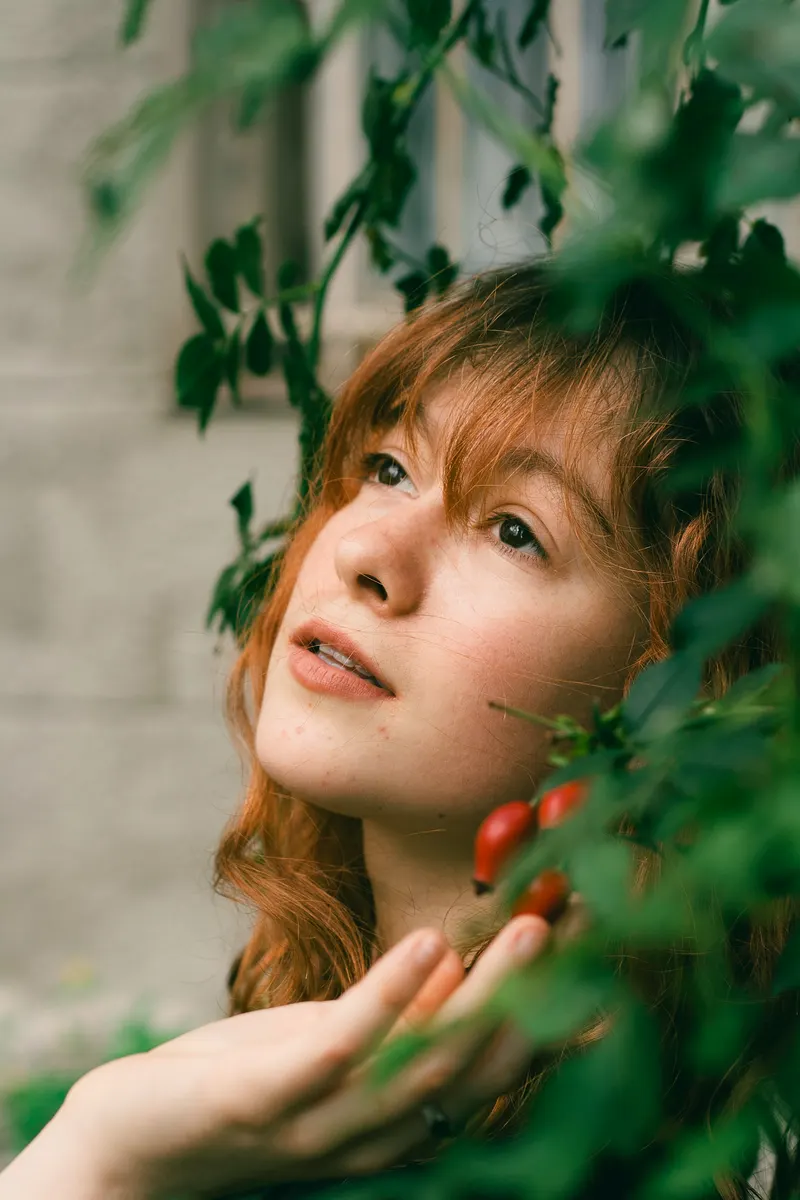 A woman with wavy copper hair tilting her head up among green leaves