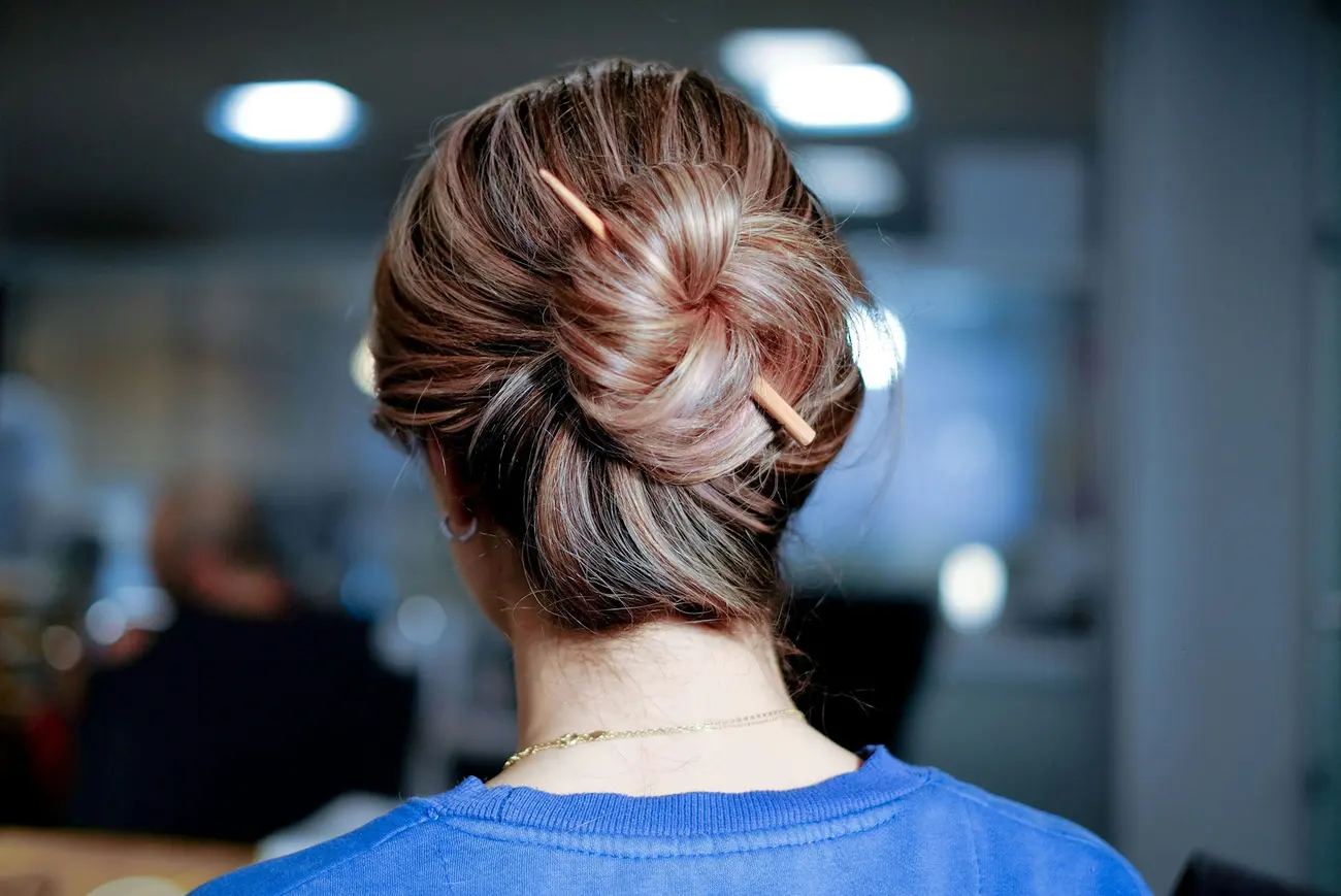 The back of a woman's head with fine blonde ribbons through light brown hair, pinned in a loose twist