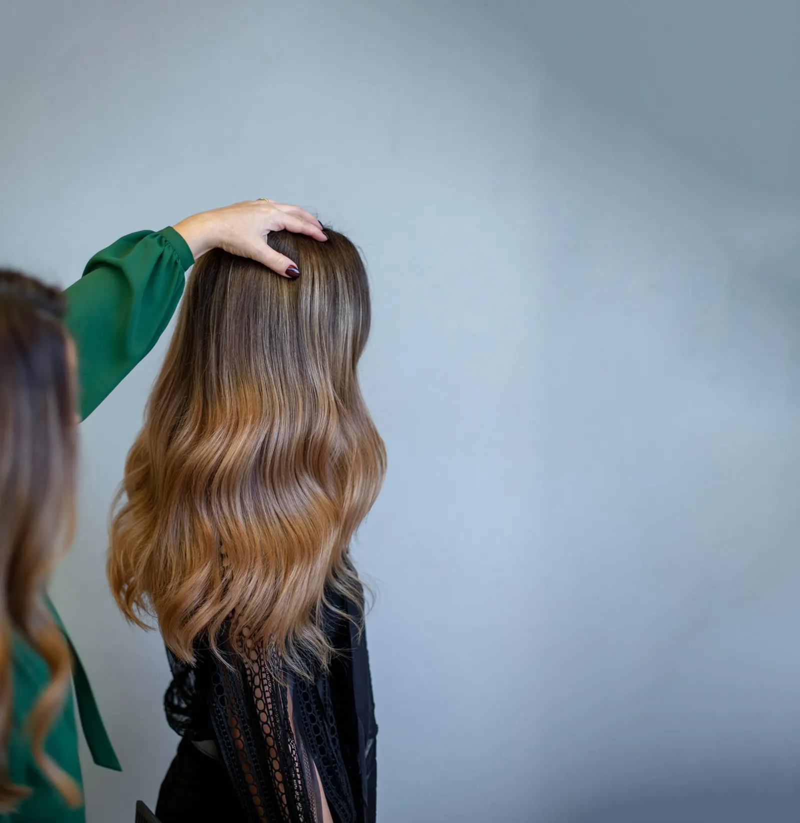 A stylist's hand lifting long, softly waved brunette hair that melts into caramel ends