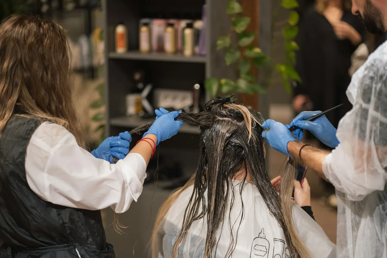 Two colourists in gloves sectioning and painting a client's hair at the colour station