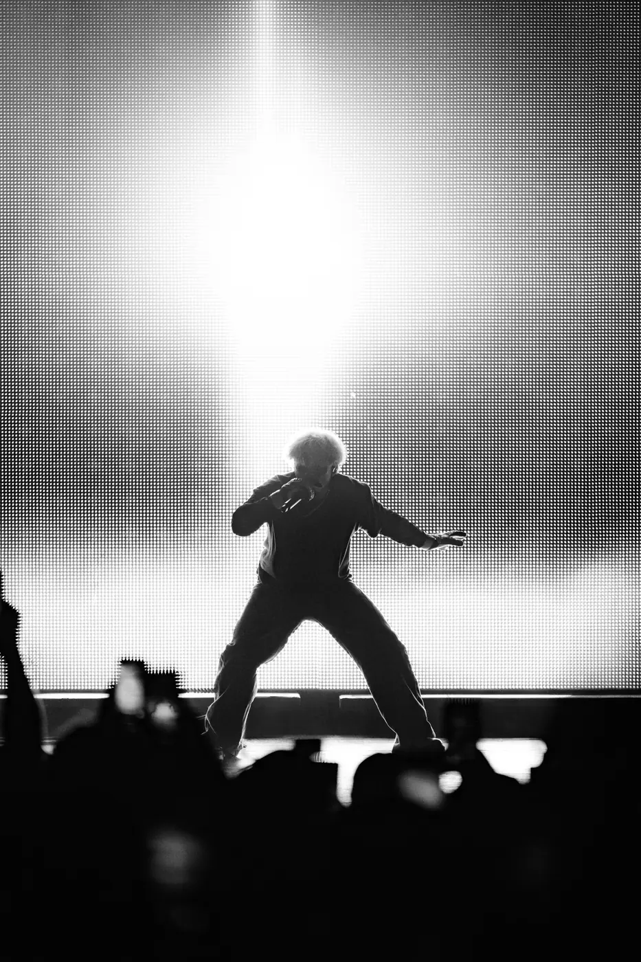 A performer mid-movement in front of a blinding white stage light, phones raised in the crowd