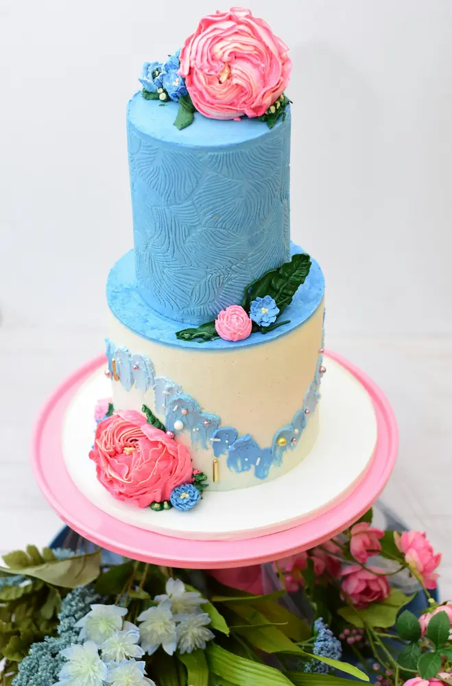 A tall two-tier cake in cornflower blue and cream with a pink peony on top