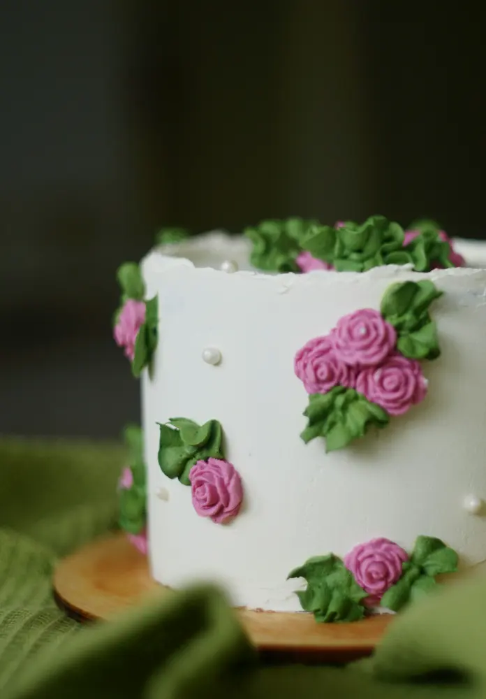 A white buttercream cake with piped pink roses and green leaves around its sides