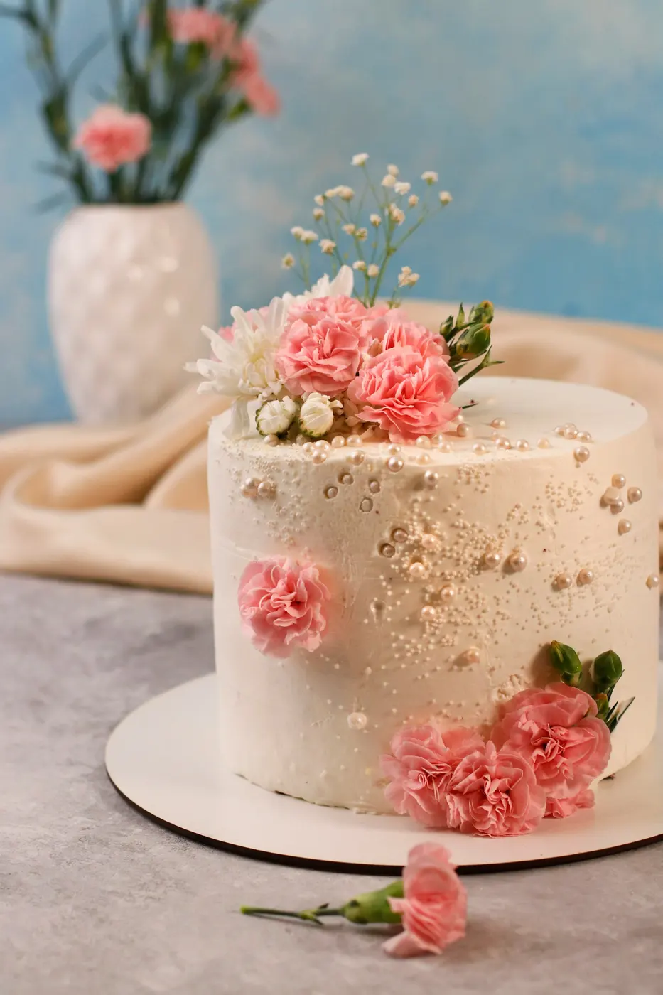 A cream buttercream cake scattered with pink flowers and gold flecks, on a cake board