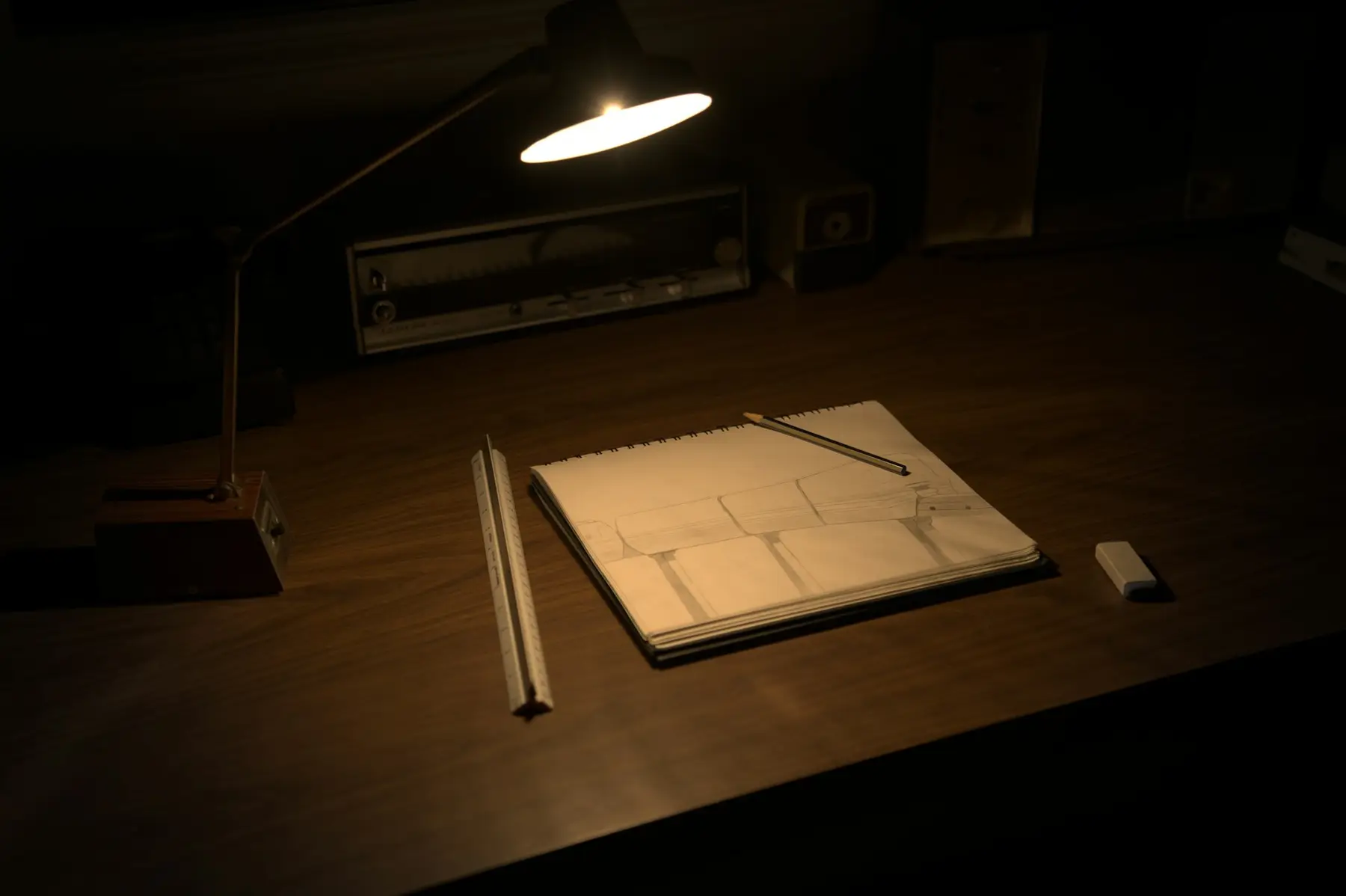 A desk at night under a single lamp, with a sketchbook, a ruler and pencils