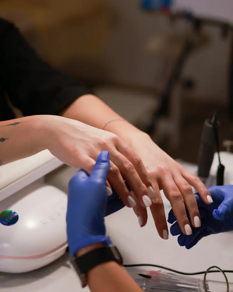 A nail tech in blue gloves finishing a client's manicure under the lamp