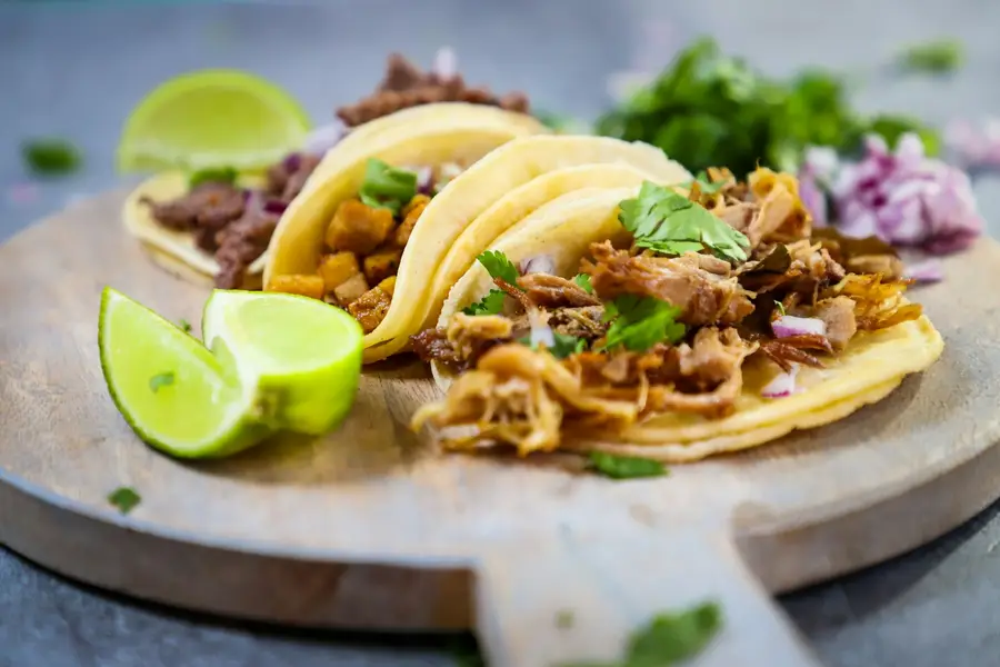 Three pork tacos on a wooden board with lime halves and cilantro