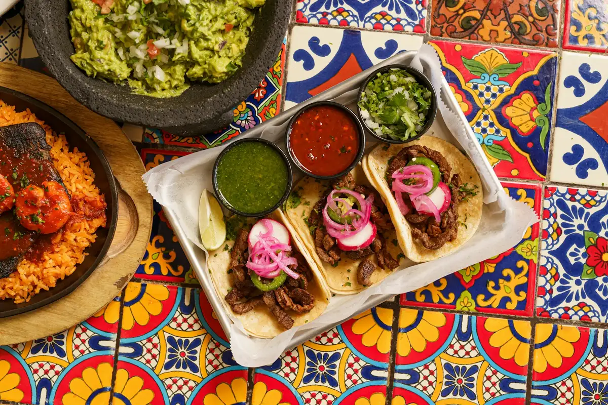 Tacos with pickled onion, bowls of salsa verde and roja, guacamole and rice on a painted Talavera tile table
