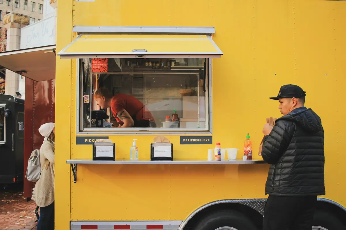 A man in a black puffer jacket waiting at the service window of a yellow food truck