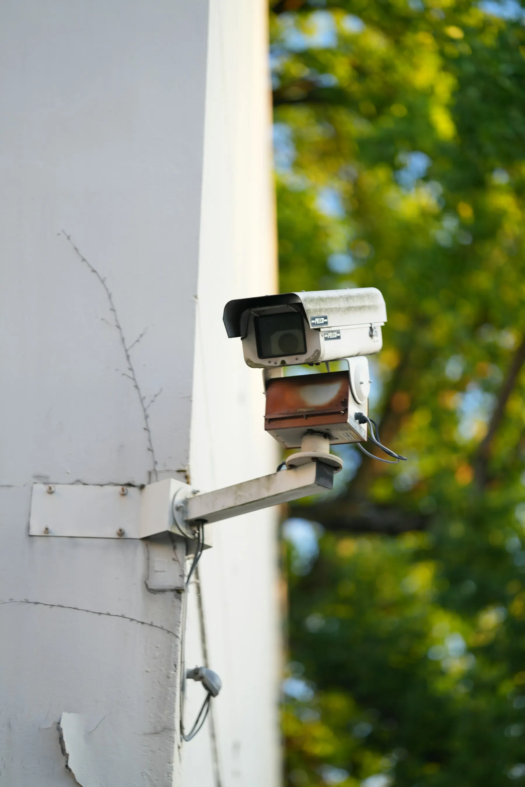A white security camera on a bracket on a pale wall, trees behind