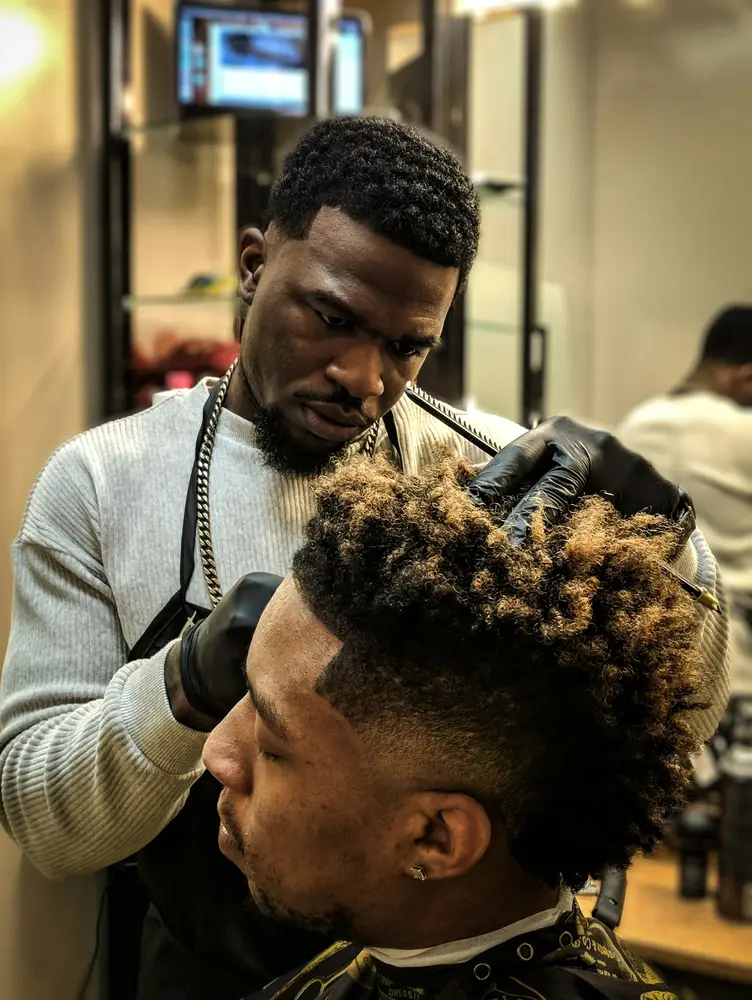 A barber in a light jacket working clippers through a client’s curly hair