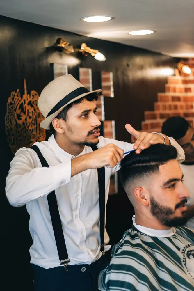 A barber in a flat cap and braces trimming a seated client’s hair