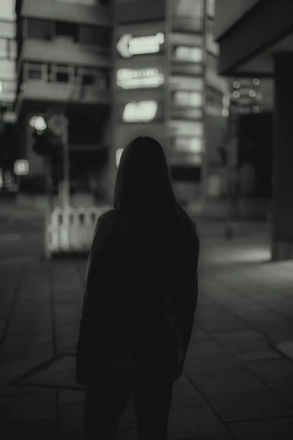 A figure in a dark hoodie stands on an empty pavement under shop signs