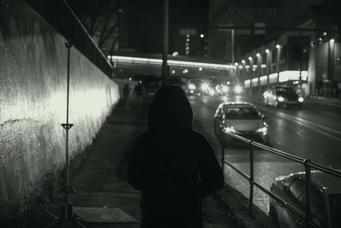 Someone in a hood walks beside a floodlit wall as cars pass at night